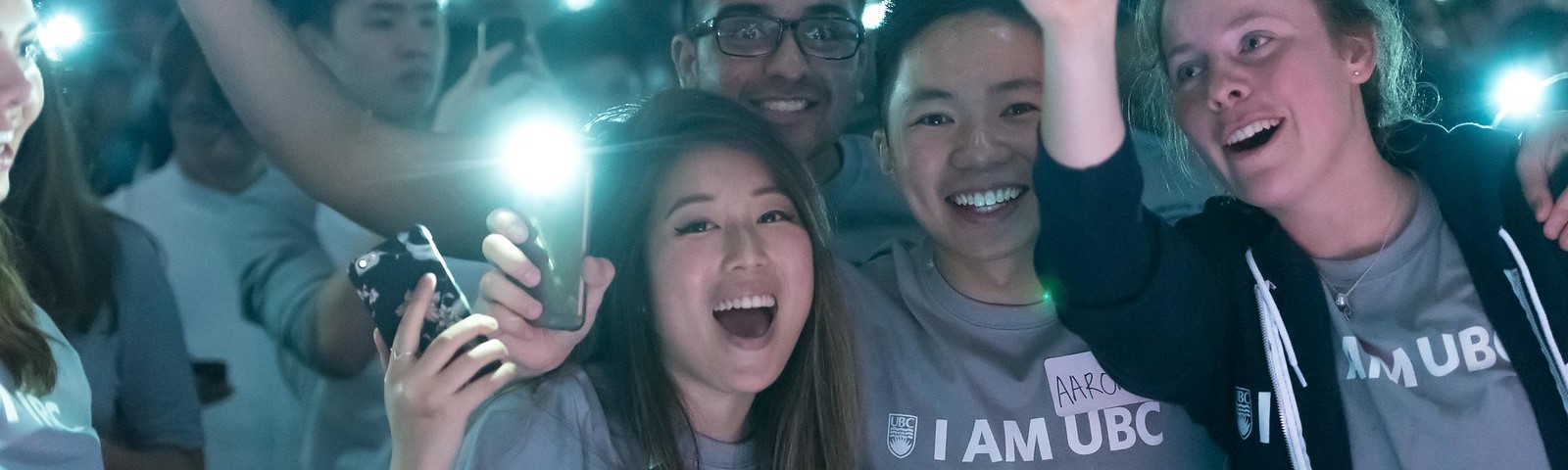 Students in matching "I am UBC" shirts cheering and using phone flashlights as celebratory communal lights