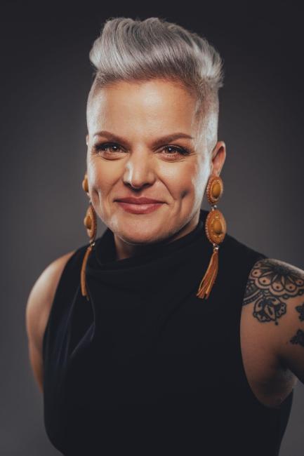 Woman with grey side-cropped hair, long leather earrings and arm tattoo