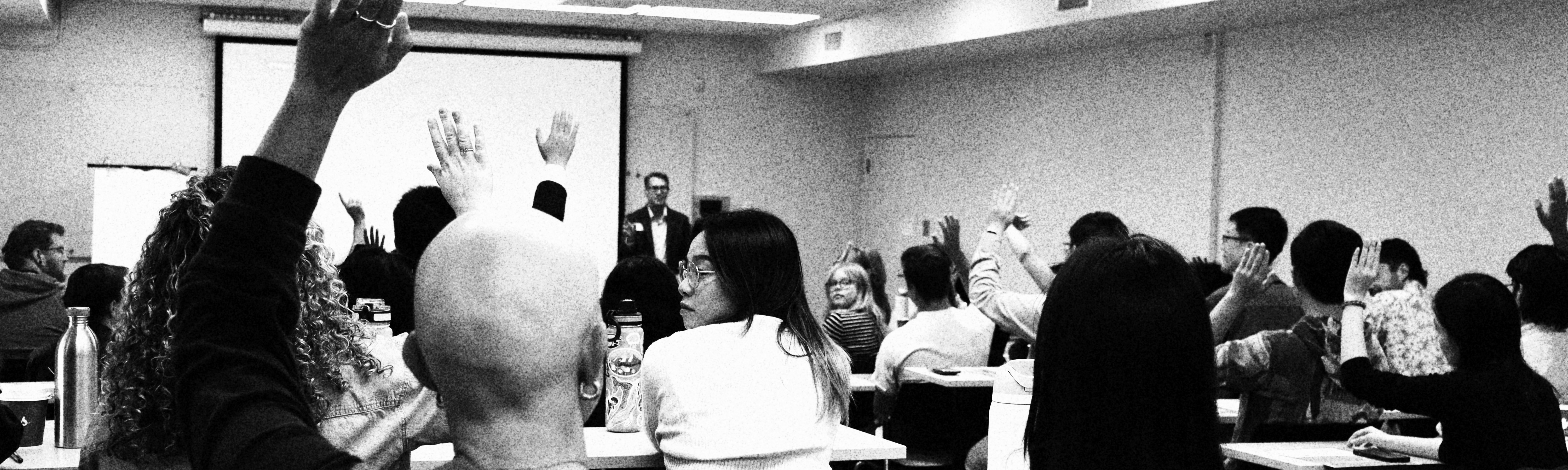 A man in a suit and glasses in front of a crowded class, more than half of which have their hands up