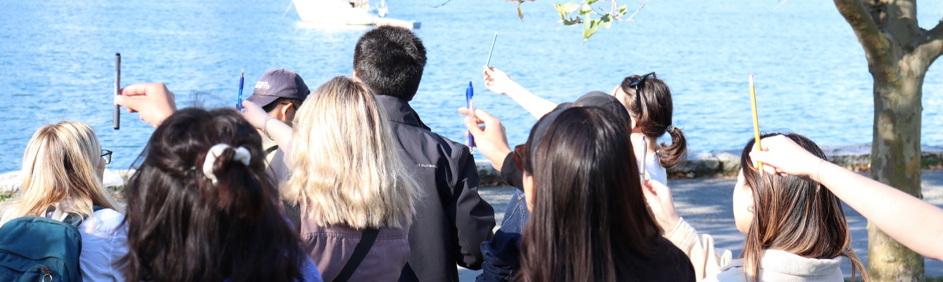 Several students hold pencils in front of their faces in front of a sunny bay