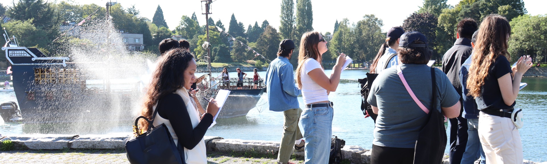 Several students sketch in front of a bay, while a parody of a pirate ship approaches them with water cannons firing free