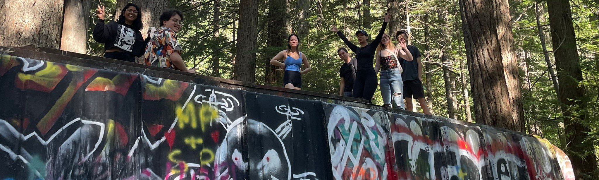 Several students posing atop an old train car covered in graffiti, in the woods