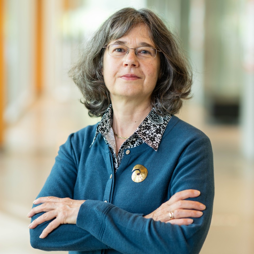 Woman in blue cardigan with gold spiral broach, arms folded, smiling slightly
