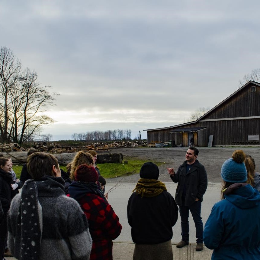 An Indigenous instructor in front of a Musqueam longhouse addressing students
