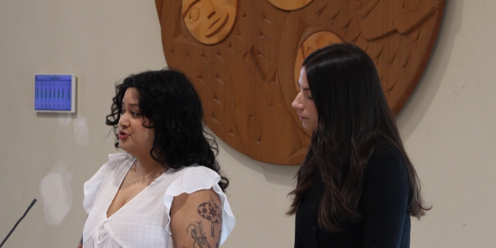 Women presenting beside a Musqueam symbol