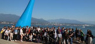 A cluster of students posing by a long blue conical sculpture, by shore and mountain range