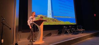 A PhD student giving a talk at a lectern, her slide showing a lighthouse