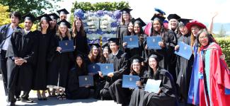 Cluster of grads in sunshine, posing around graduation poster