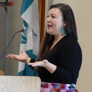 Indigenous woman speaking at podium beside Musqueam Indian Band flag