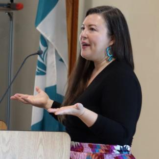 Indigenous woman speaking at podium beside Musqueam Indian Band flag