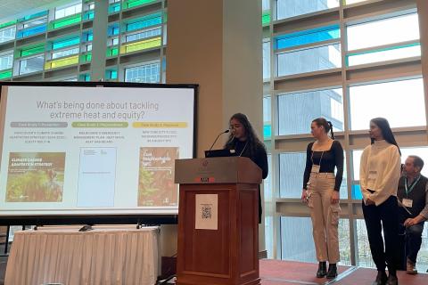 A student at a lectern presenting, with two students standing at her side