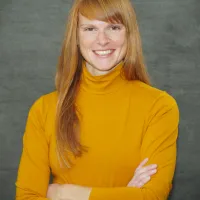 Maggie in orange turtleneck, smiling with arms folded