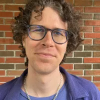 Person in front of brick wall, in purple shirt and necklace